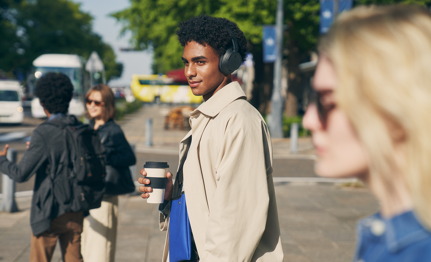 Image of a person in a town wearing a black pair of Sony WH-CH720N headphones holding a cup of coffee.